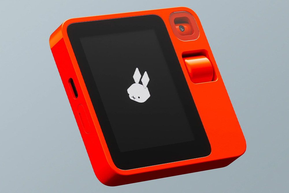 Ein leuchtend oranges KI-Gerät Rabbit R1 mit einem kleinen Bildschirm, auf dem das Logo eines stilisierten Hasen angezeigt wird, sowie einer rotierenden Kamera und einem Navigationsrad an der Seite.