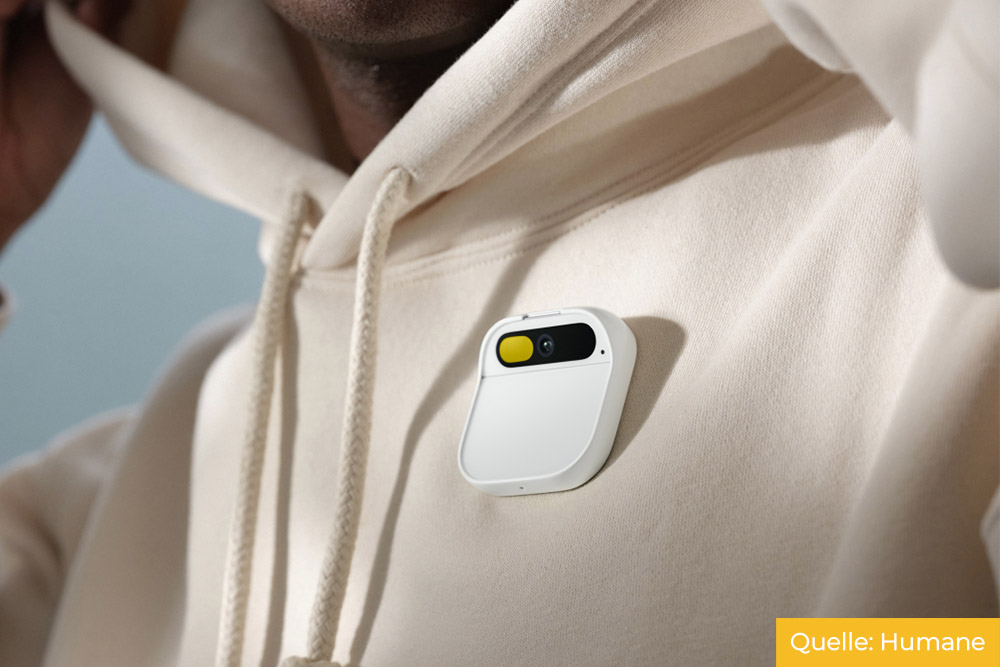 Eine Person in einem hellbeigen Hoodie trägt Humane's Ai Pin, ein weißes, rechteckiges Wearable mit einer gelb-schwarzen Oberfläche.